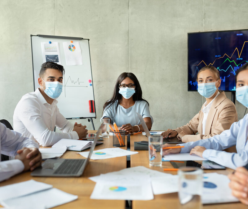 Safety At Work. Diverse Business People In Protective Medical Face Masks Having Corporate Meeting In Office, Multiethnic Colleagues Sitting At Desk And Looking At Camera, Working During Pandemic